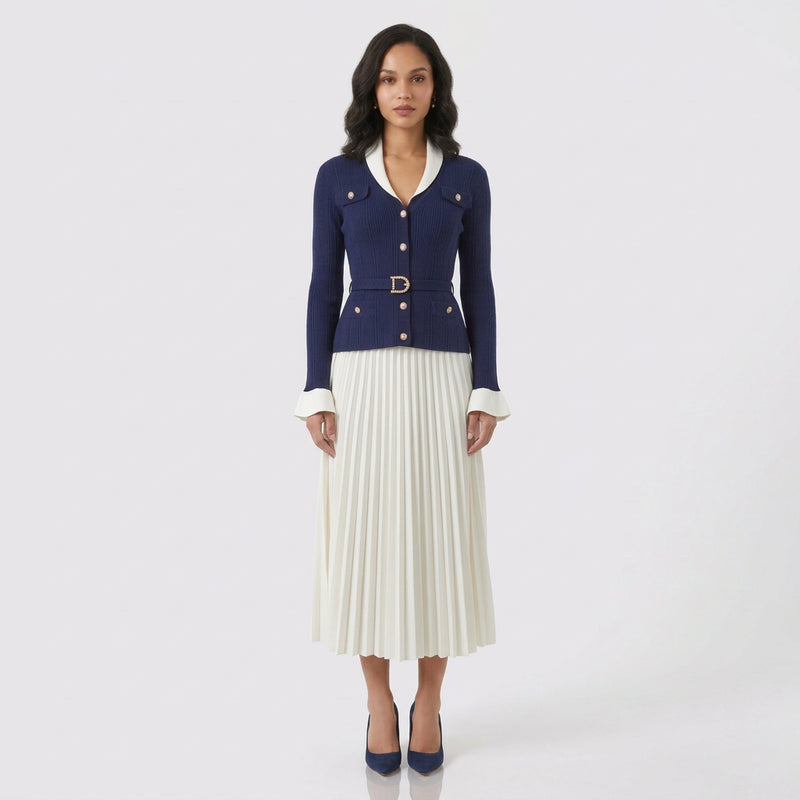 A model stands against a light gray studio background wearing a navy blue cropped cardigan with white trim and a matching belt, paired with a white pleated midi skirt and navy blue heels.