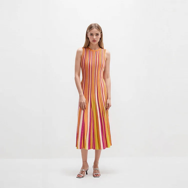 A woman stands against a plain white studio background wearing a sleeveless midi dress with vertical pink, orange, yellow, and white stripes, paired with metallic heeled sandals.