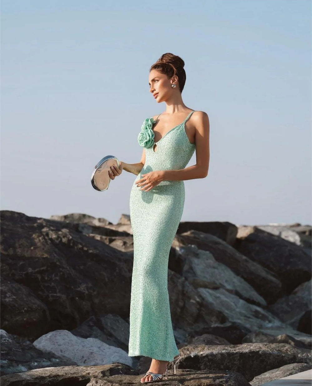 Woman in a light green evening gown standing on rocks with a clear sky background