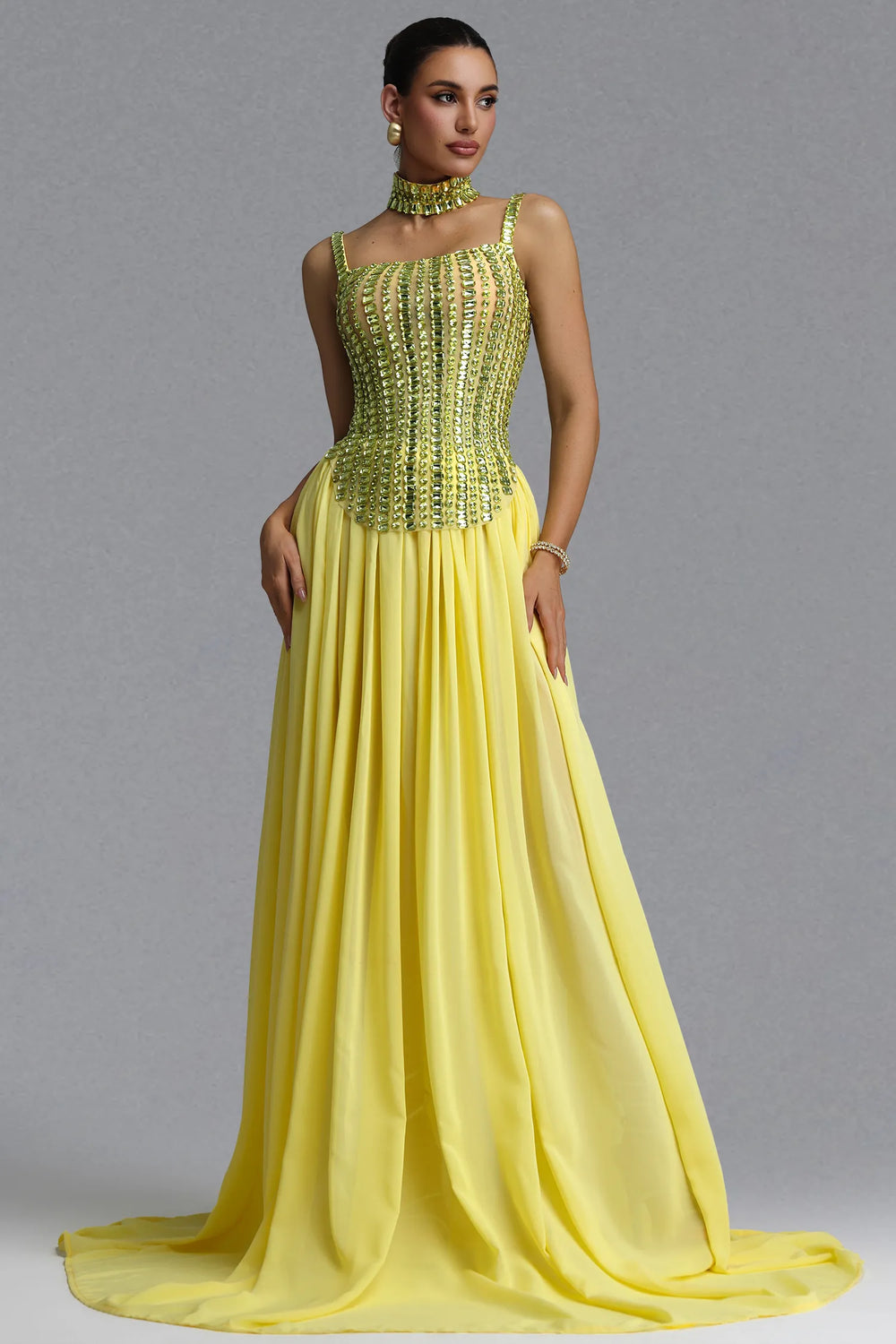 A woman models a floor-length yellow gown with a heavily embellished beaded corset-style bodice and a flowing chiffon skirt, photographed against a plain gray studio background.