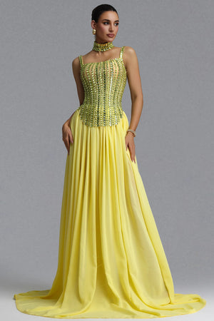 A woman models a floor-length yellow gown with a heavily embellished beaded corset-style bodice and a flowing chiffon skirt, photographed against a plain gray studio background.