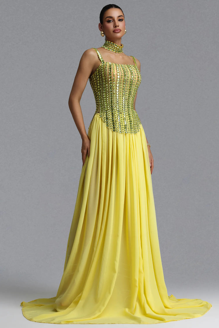A woman models a floor-length yellow gown with a heavily embellished beaded corset-style bodice and a flowing chiffon skirt, photographed against a plain gray studio background.