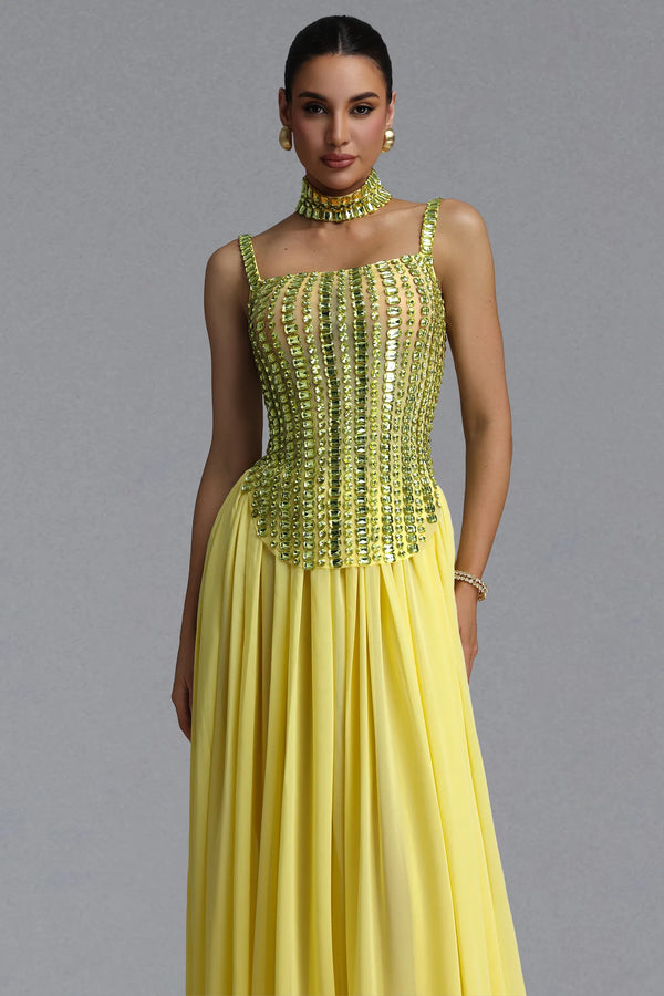 A woman modeling a floor-length yellow gown with a heavily embellished corset-style bodice in metallic yellow and green sequins, paired with a matching choker necklace and bracelet, against a plain gray studio background.