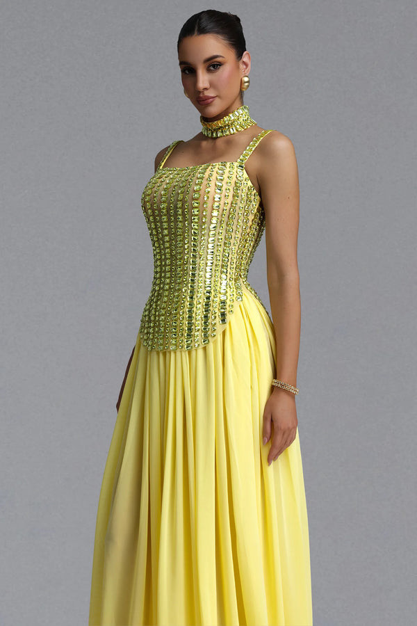 A woman modeling a yellow formal gown with a heavily embellished gold sequined corset bodice, a matching gold choker, and a flowing chiffon skirt, set against a plain gray studio background.