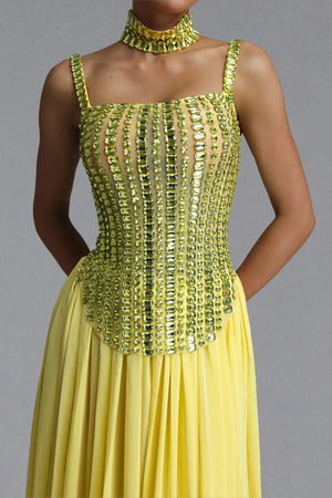 A woman wearing a bright yellow formal dress with a heavily embellished corset-style bodice covered in vertical rows of shiny gold and green sequins, paired with a matching gold choker necklace, against a plain gray studio background.