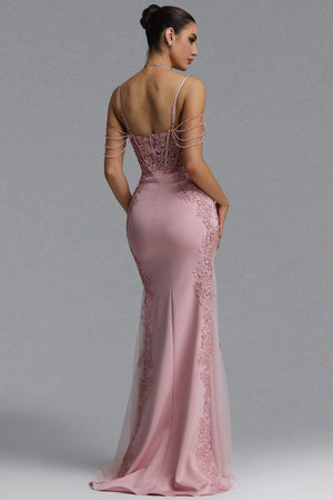A woman modeling a floor-length dusty pink evening gown from the back, featuring a sheer lace-embellished back bodice with thin shoulder straps and a flared tulle skirt, against a plain gray studio background.