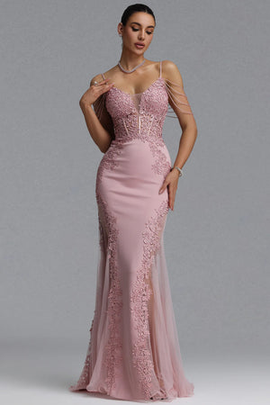 A woman models a floor-length pink evening gown with a fitted bodice, lace applique, thin shoulder straps, and a sheer tulle skirt against a plain gray studio background.