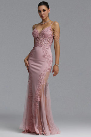 A woman modeling a floor-length dusty pink evening gown with lace applique, sheer mesh panels, and thin shoulder straps against a plain gray studio background.