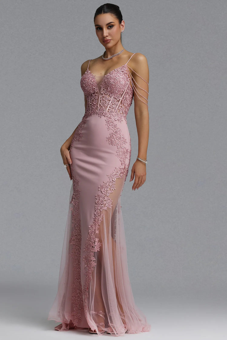 A woman models a floor-length pink evening gown with a fitted bodice, lace applique, thin shoulder straps, and a sheer tulle skirt against a plain gray studio background.