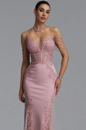 A woman modeling a floor-length pink evening gown with thin straps, a structured lace bodice, sheer mesh panels, and delicate chain draping across the shoulders, photographed against a plain gray studio background.