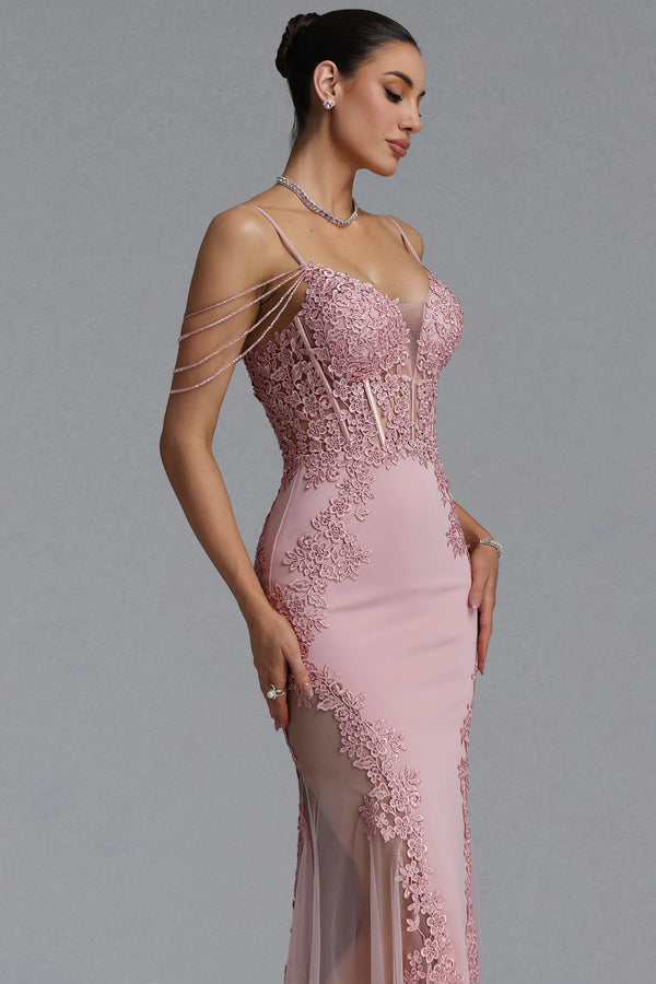 A woman modeling a formal evening gown in dusty pink with floral lace applique, thin straps, and delicate chain draping across the shoulders, against a plain gray studio background.