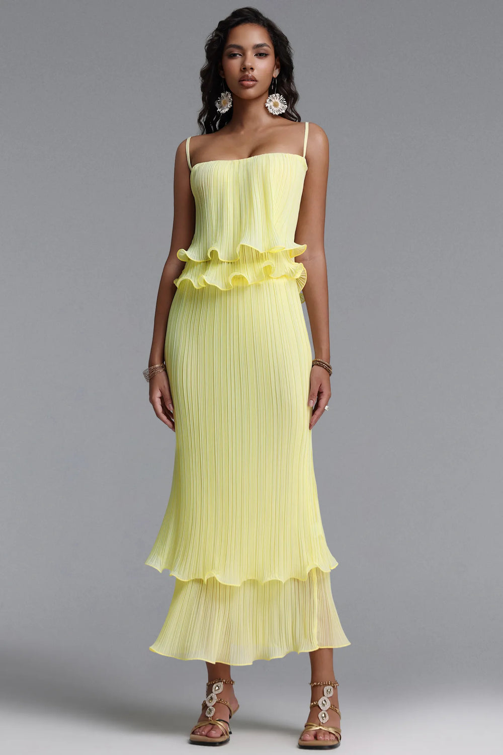 A woman modeling a pale yellow pleated midi dress with spaghetti straps, a ruffled peplum waist, and a tiered skirt, paired with large statement earrings and strappy sandals, against a solid gray studio background.