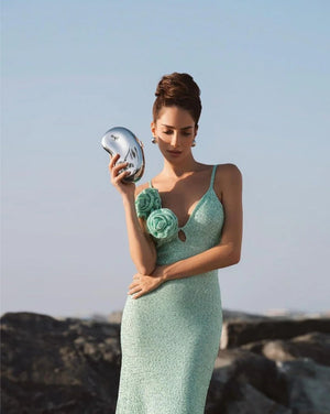 Woman in a light green dress holding a silver mirror on a rocky beach.