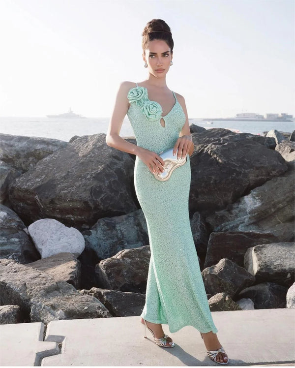 Woman in a light green evening gown with floral details standing on rocks by the water.