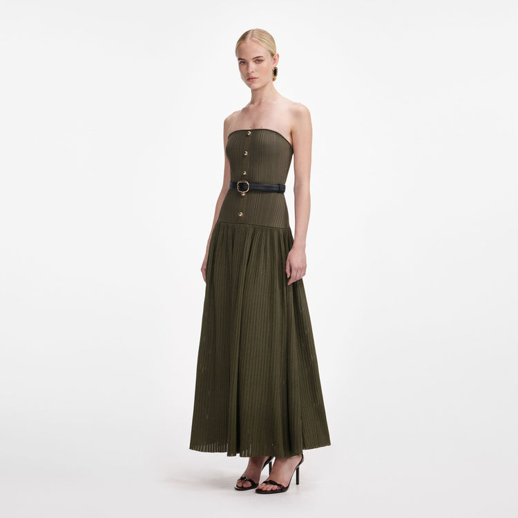 A woman modeling a strapless olive green pleated maxi dress with a black waist belt and gold-tone buckle, set against a plain white studio background.