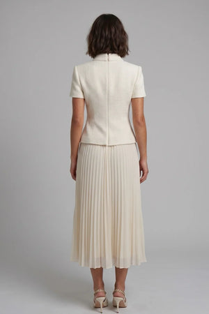 A woman viewed from the back wearing a cream-colored short-sleeve jacket and matching pleated midi skirt, paired with beige high-heeled sandals, against a solid gray studio background.