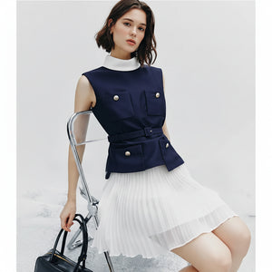 Woman wearing a navy sleeveless top with white pleated skirt, holding a black handbag.