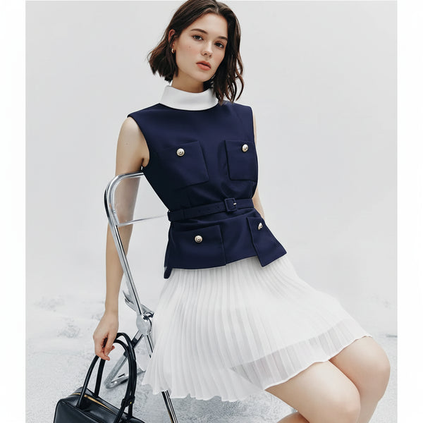 Woman wearing a navy sleeveless top with white pleated skirt, holding a black handbag.