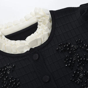 Black textured garment with a white knit scarf and black embellishments on a light gray background