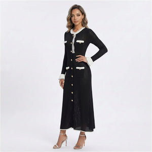 A woman modeling a long black textured dress with white lace trim at the collar and cuffs, and gold-tone buttons down the front, photographed against a light gray studio background.