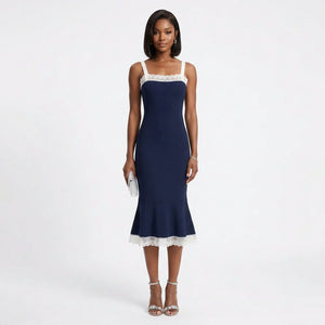 A woman modeling a navy midi dress with white lace trim at the neckline and hem, paired with silver strappy heels and holding a small white clutch, against a plain white studio background.