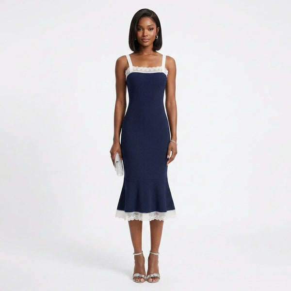 A woman modeling a navy midi dress with white lace trim at the neckline and hem, paired with silver strappy heels and holding a small white clutch, against a plain white studio background.