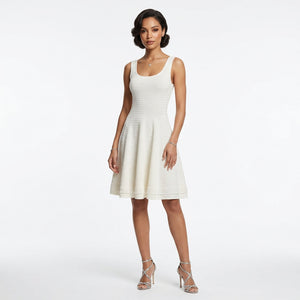 Maiwenn Scoop-Neck Knit Mini Dress — A woman modeling a sleeveless, knee-length textured dress in a warm off-white or cream color, paired with metallic silver high-heeled sandals, against a plain white studio background.