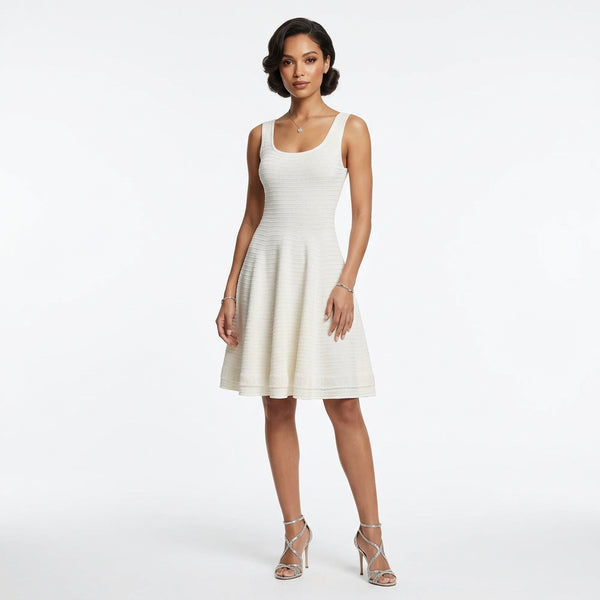 Maiwenn Scoop-Neck Knit Mini Dress — A woman modeling a sleeveless, knee-length textured dress in a warm off-white or cream color, paired with metallic silver high-heeled sandals, against a plain white studio background.