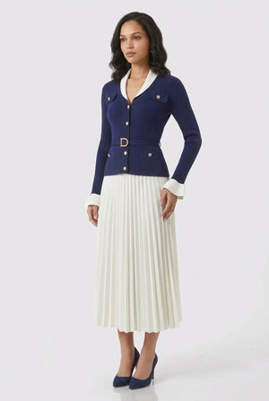 A model stands against a light gray studio background wearing a navy blue cropped cardigan with white trim and a matching belt, paired with a white pleated midi skirt and navy blue heels.