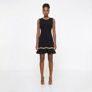 A woman modeling a sleeveless black mini dress with a ruffled hem and a thin gold trim detail at the waist, paired with gold high heels against a plain white studio background.