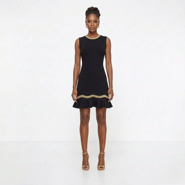 A woman modeling a sleeveless black mini dress with a ruffled hem and a thin gold trim detail at the waist, paired with gold high heels against a plain white studio background.