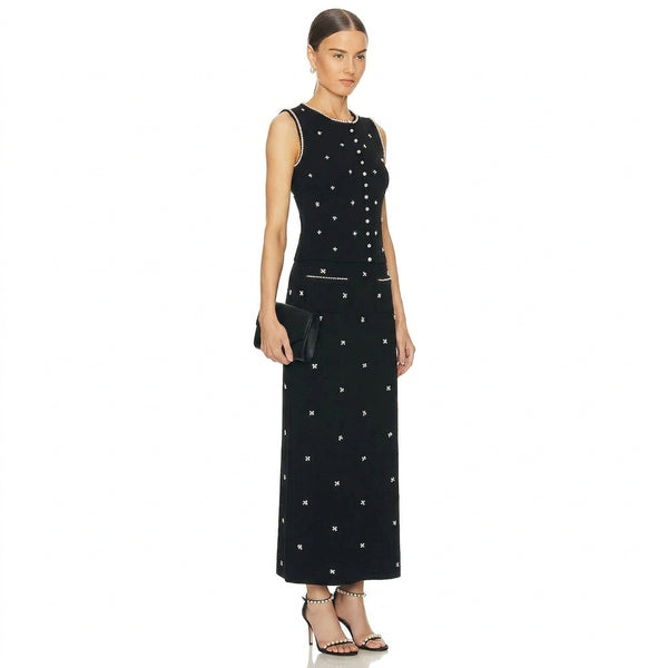 A woman modeling a sleeveless black maxi dress with white embroidered star patterns, white piping details, and a white button-up front, set against a plain white background.