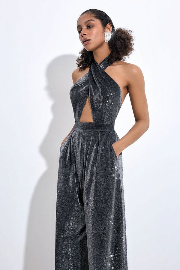 A woman modeling a sparkling dark gray halter-neck jumpsuit with a cutout bodice and wide legs, standing against a plain light background.