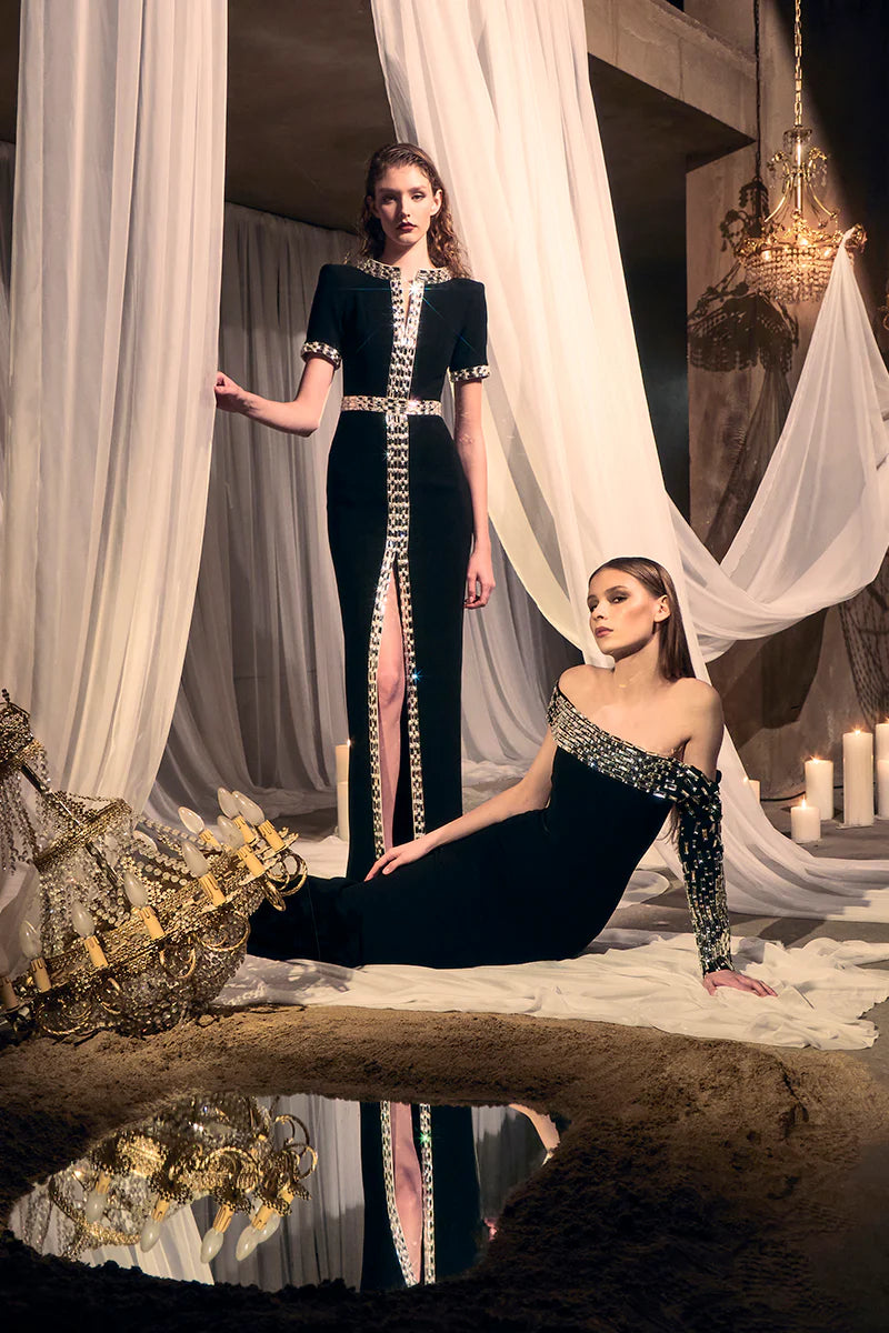 Two women in elegant black dresses with gold accents standing in a luxurious setting with white curtains and candles.