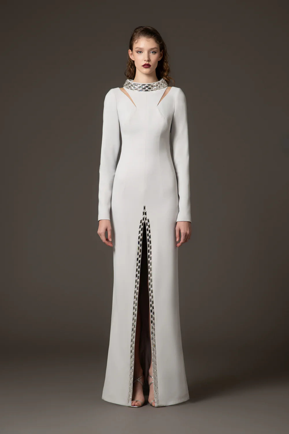 A model stands against a dark gray studio background wearing a floor-length white gown with long sleeves, a high neckline with cutout details, and a vertical slit lined with a black-and-white patterned trim.