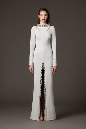 A model stands against a dark gray studio background wearing a floor-length white gown with long sleeves, a high neckline with cutout details, and a vertical slit lined with a black-and-white patterned trim.