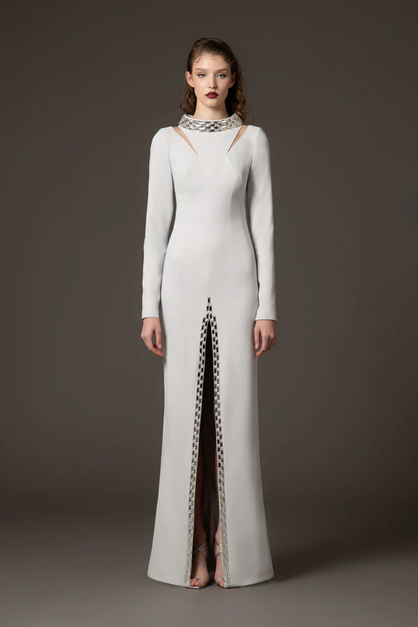 A model stands against a dark gray studio background wearing a floor-length white gown with long sleeves, a high neckline with cutout details, and a vertical slit lined with a black-and-white patterned trim.