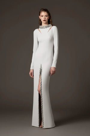 A model stands against a dark gray studio background wearing a floor-length white gown with long sleeves, a high neckline with cutout details, and a vertical slit lined with a black-and-white patterned chain.