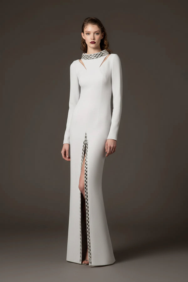 A model stands against a dark gray studio background wearing a floor-length white gown with long sleeves, a high neckline with cutout details, and a vertical slit lined with a black-and-white patterned chain.