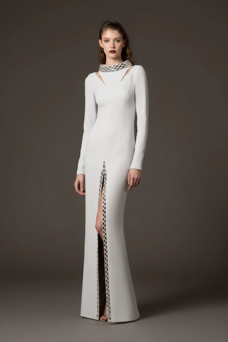 A model stands against a dark gray studio background wearing a floor-length white gown with long sleeves, a high neckline with cutout details, and a vertical slit lined with a black-and-white patterned trim.