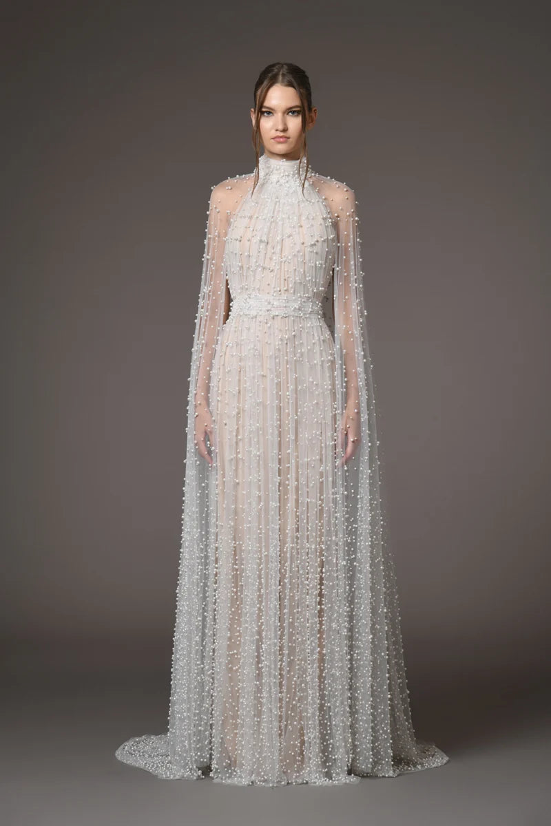 A model wearing a floor-length, high-neck white gown with sheer draped cape sleeves and dense beaded vertical texture, photographed against a dark gray studio background.
