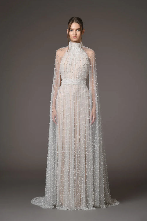 A model wearing a floor-length, high-neck white gown with sheer draped cape sleeves and dense beaded vertical texture, photographed against a dark gray studio background.