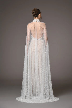 A model shown from the back wearing a floor-length white gown with a sheer high neck, long draped cape sleeves, and an open back, photographed against a plain taupe studio background.
