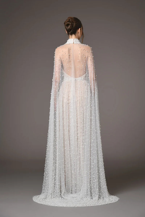 A model shown from the back wearing a floor-length white gown with a sheer high neck, long draped cape sleeves, and an open back, photographed against a plain taupe studio background.