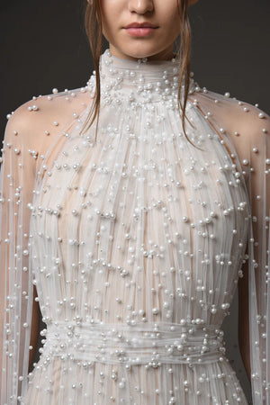A close-up of a model wearing a high-neck, sheer champagne-colored dress heavily embellished with white pearls and beadwork, set against a dark gray studio background.