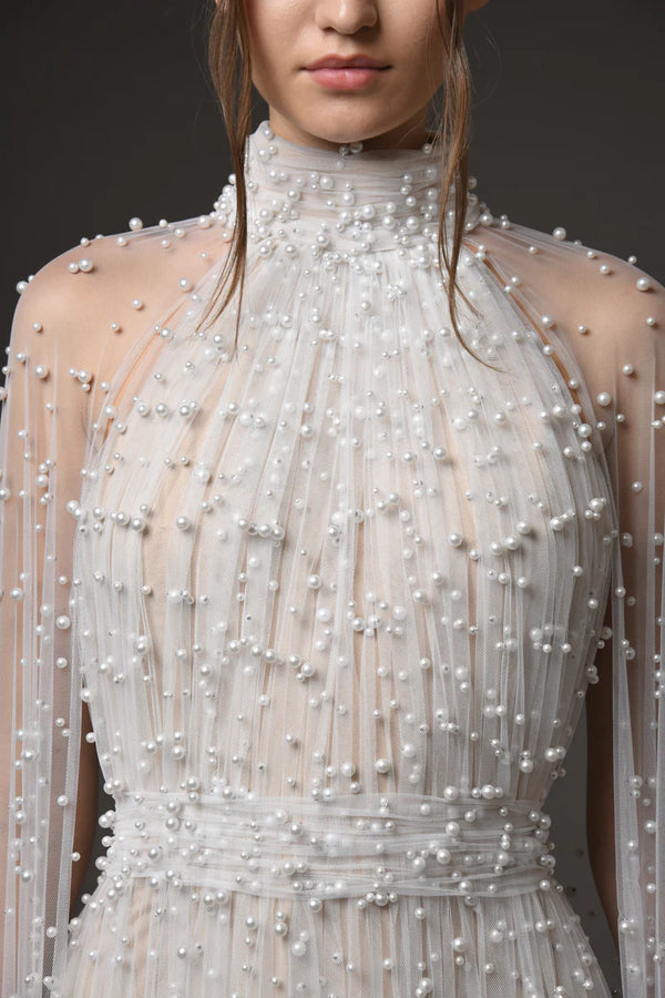A close-up of a model wearing a high-neck, sheer champagne-colored dress heavily embellished with white pearls and beadwork, set against a dark gray studio background.
