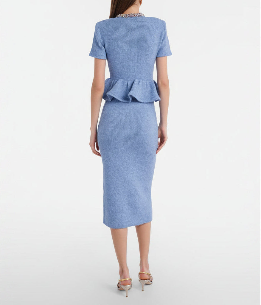 A woman models a light blue tweed two-piece set consisting of a short-sleeve peplum button-front jacket and a matching knee-length pencil skirt, styled with metallic pointed-toe heels and a small metallic clutch against a plain light gray studio background.