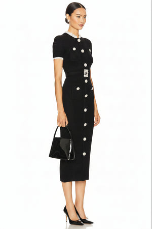 A model wearing a black short-sleeve midi dress with white buttons and a decorative belt buckle, holding a black handbag against a plain white background.