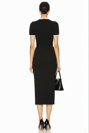 A model wearing a black short-sleeve midi dress with white buttons and a decorative belt buckle, holding a black handbag against a plain white background.
