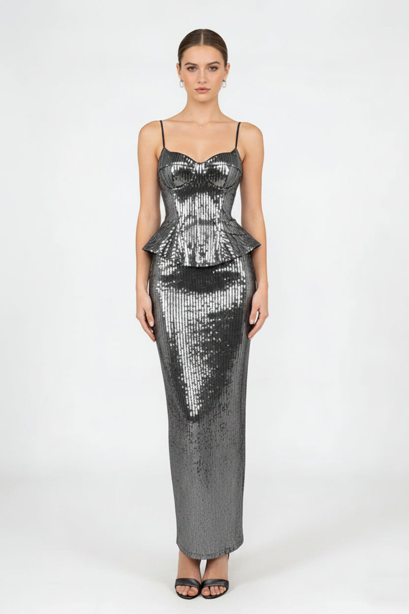 A woman modeling a floor-length, bodycon sequined dress in metallic silver with thin spaghetti straps and a sweetheart neckline, paired with a matching sequined clutch bag and black sandals, against a plain light gray studio background.
